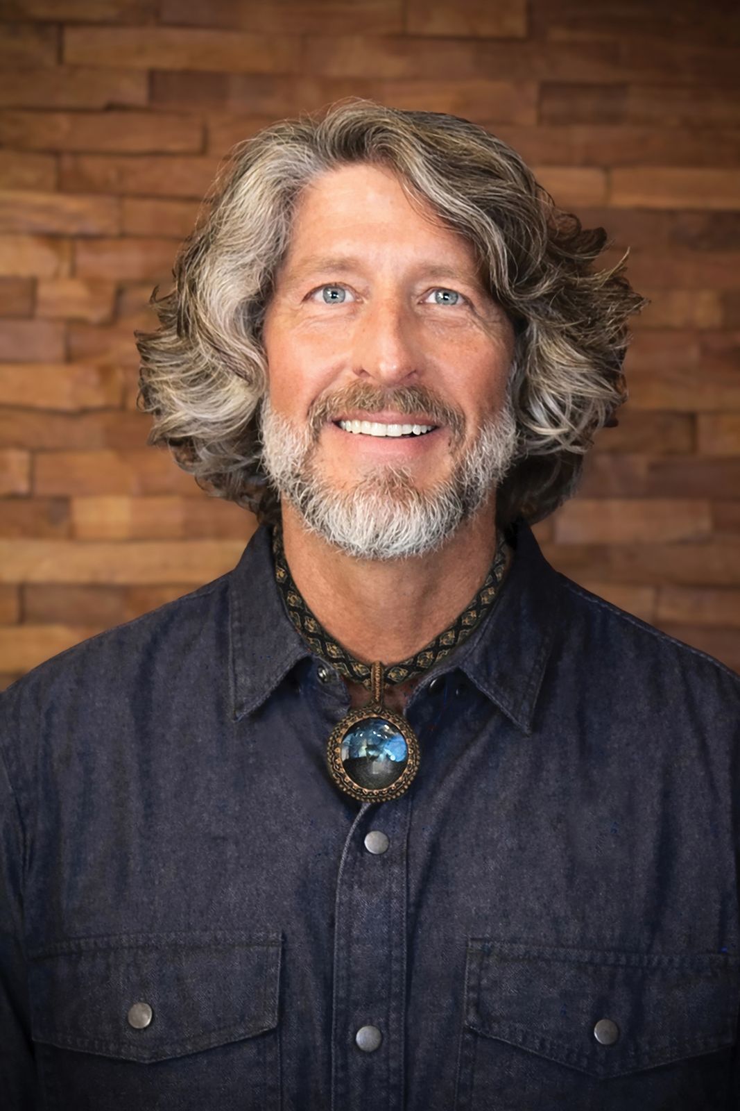 Portrait of Marc Buckley smiling in a dark shirt and pendant, against a brick wall